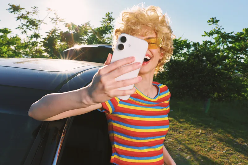Casual woman takes a cheerful selfie outdoors beside a car, wearing a striped colorful shirt, capturing a spontaneous joy moment with a retro film color vibe.