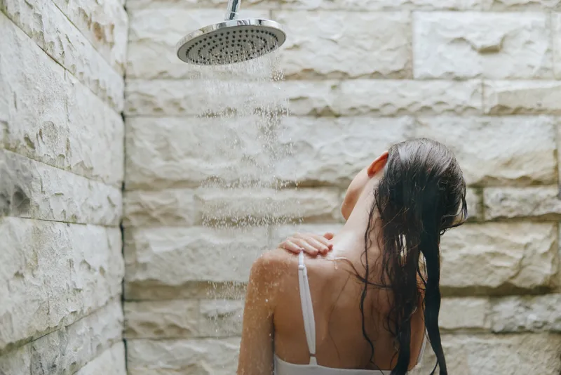 woman taking shower under outdoor rain shower with stone wall background, wet long hair, white strap top, natural spa moment, refreshing water droplets