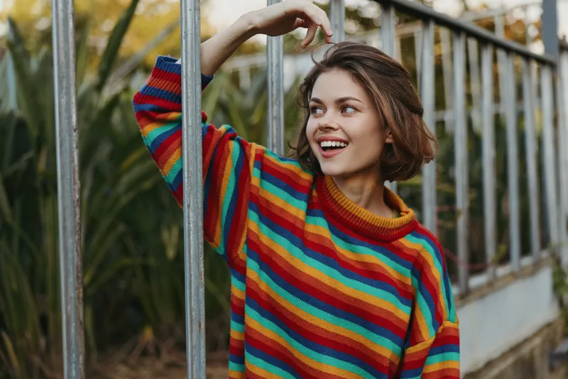 Young woman in her twenties wearing a colorful striped sweater, smiling and leaning on a metal fence outdoors amid tropical plants, casual fashion portrait for lifestyle or travel.