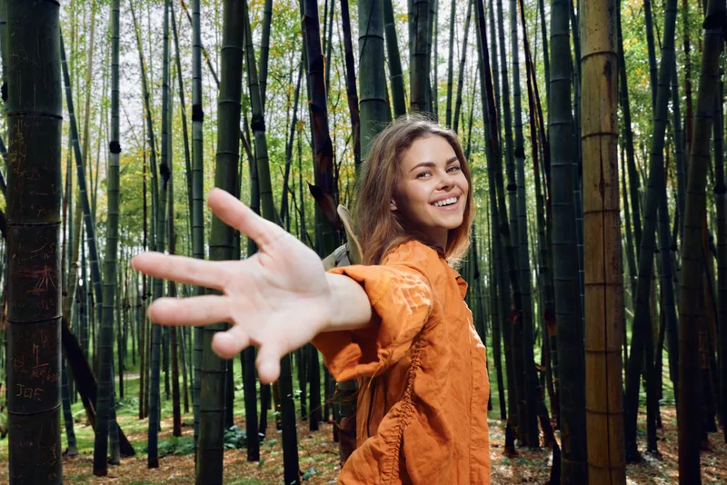 Woman smile bamboo forest hand reaching toward camera, portrait of happy young woman outdoors in nature wearing orange jacket, looking back and inviting viewer on a woodland walk.