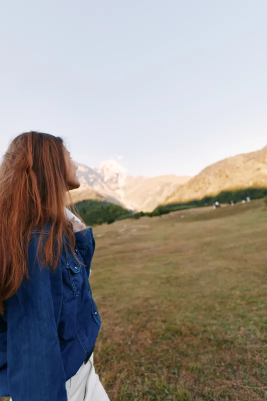Woman redhead mountains meadow denim outdoors, profile portrait of young woman standing in grassy field, looking toward distant peaks at sunrise for travel, hiking and peaceful nature escape.