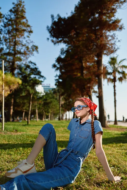 Lifestyle woman wearing modern boho-western denim outfit and sunglasses, relaxing on grass in sunny park with film color effect and street style vibe.