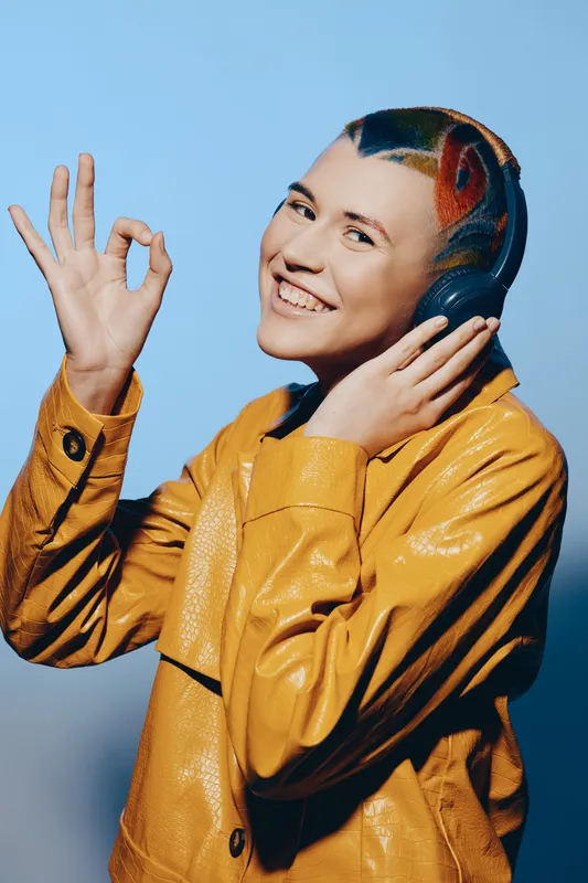 smiling woman with colorful hair listening to music in headphones wearing yellow jacket expressing approval sign against blue background