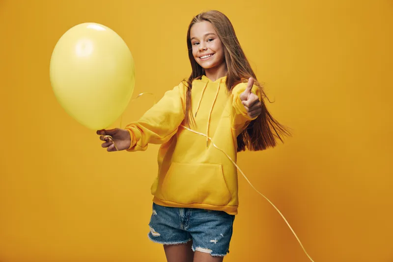 Happy girl holding yellow balloon and showing thumbs up isolated on yellow background, smiling child in casual clothes with long hair for celebration or fun.