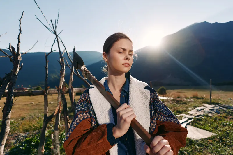 woman outdoors mountains sunrise rural stick jacket nature A young woman in a warm jacket holds a wooden stick in a sunlit field, eyes closed and serene against distant mountain peaks.