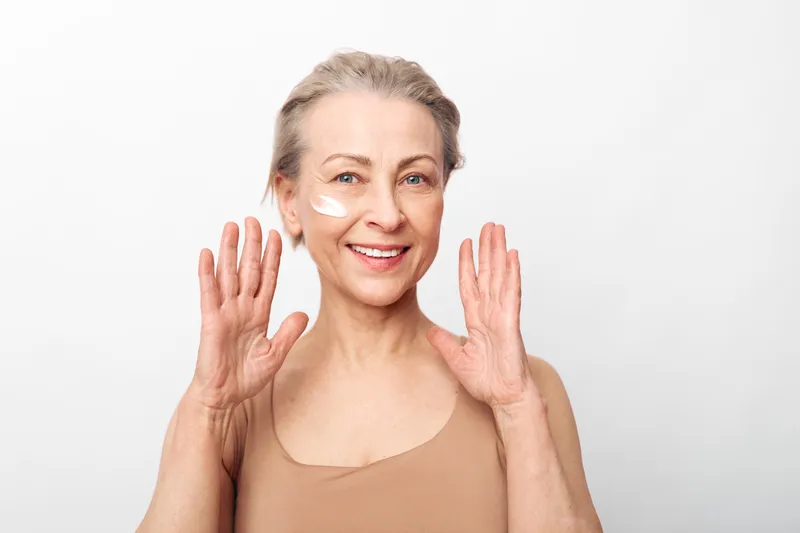 Smiling mature woman with facial cream on cheek raises hands in joyful gesture. Isolated on white background, concept of skincare, self-care, beauty routine, and natural aging confidence.