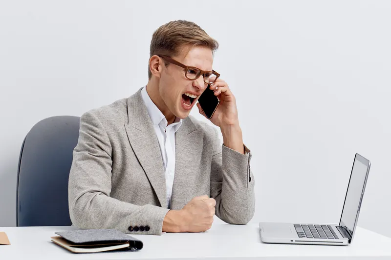 Confident businessman in gray suit and white shirt shouting while talking on mobile phone at modern office desk. Business communication concept, workplace stress, corporate environment, professional a