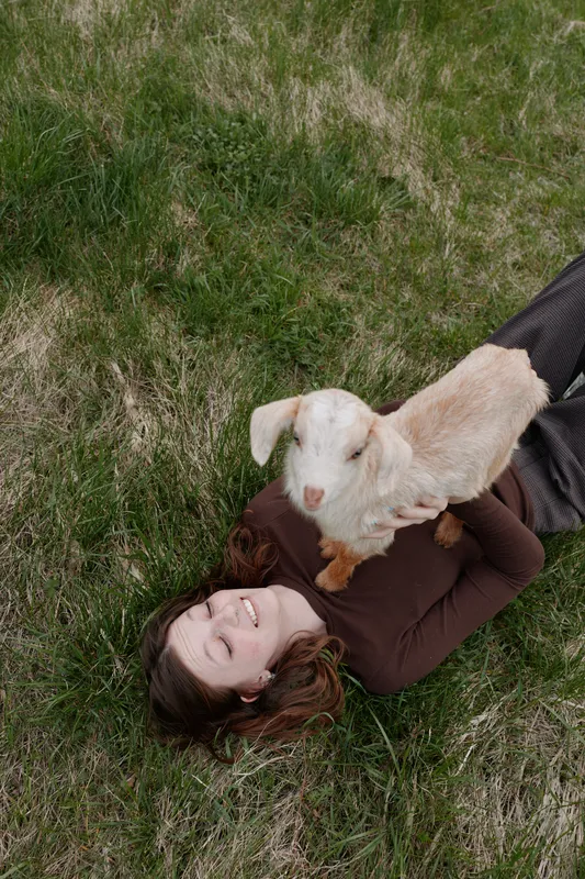 Relaxed woman lying on grass with a playful goat kid perched on her chest, outdoor lifestyle moment highlighting affection and cuddle, nature and companionship in a gentle, joyful scene.