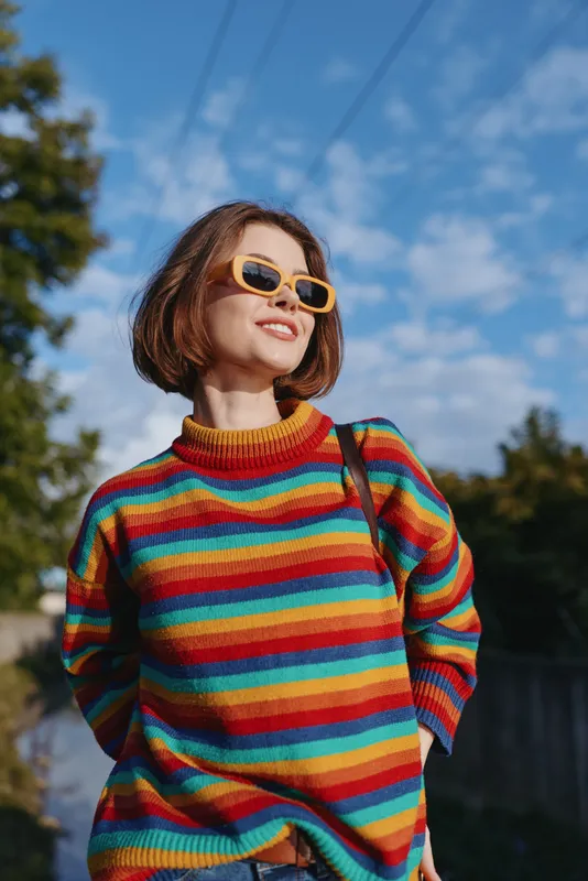 Woman in rainbow sweater wearing yellow sunglasses, smiling in outdoor portrait under blue sky and warm golden hour light. Casual fashion, short bob hairstyle and confident happy expression.
