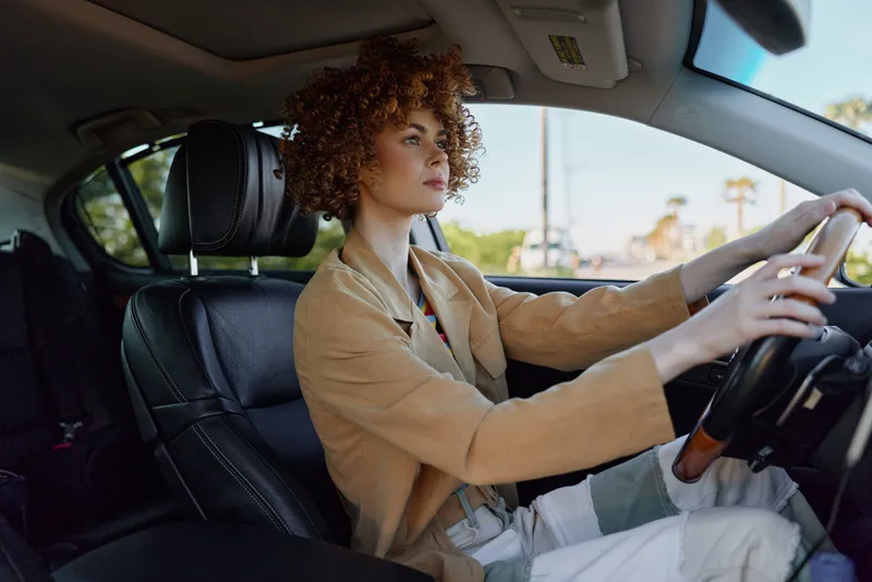 Young woman with curly hair drives a car in a sunlit street, calm focus on her face as hands grip the steering wheel, a candid moment of daily independence.