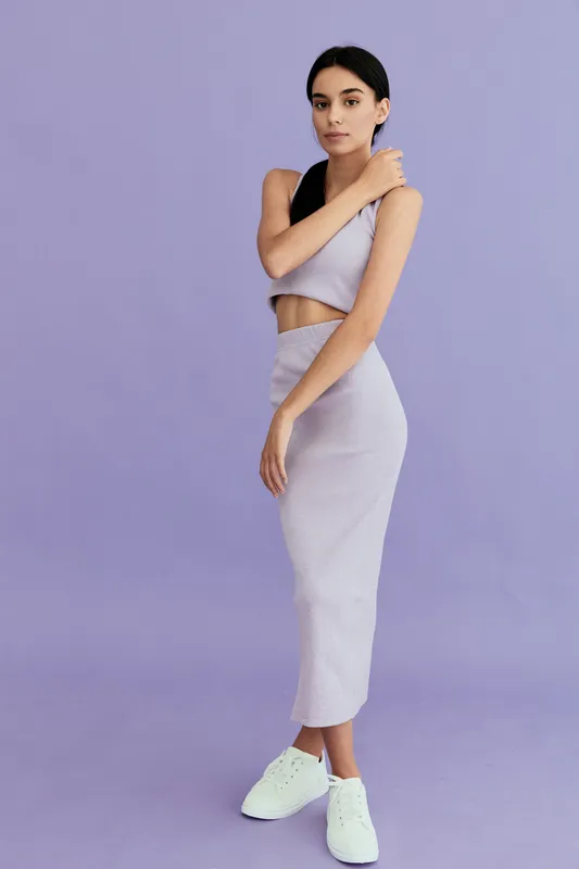 Young woman wearing a stylish lilac two-piece outfit and white sneakers stands confidently against a solid lavender background, showcasing modern fashion and minimalism.