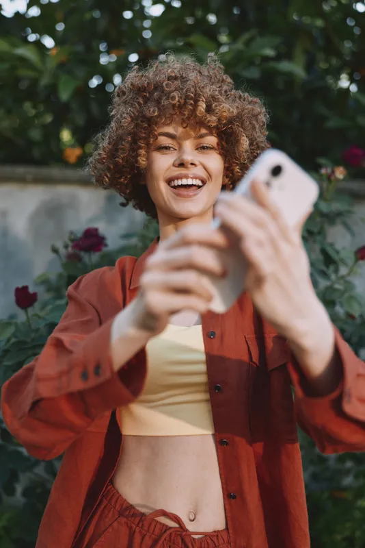 Smiling woman with curly hair taking a selfie in a sunny garden, wearing a red shirt and pale yellow top. Casual lifestyle, outdoor joy, candid portrait, vibrant mood