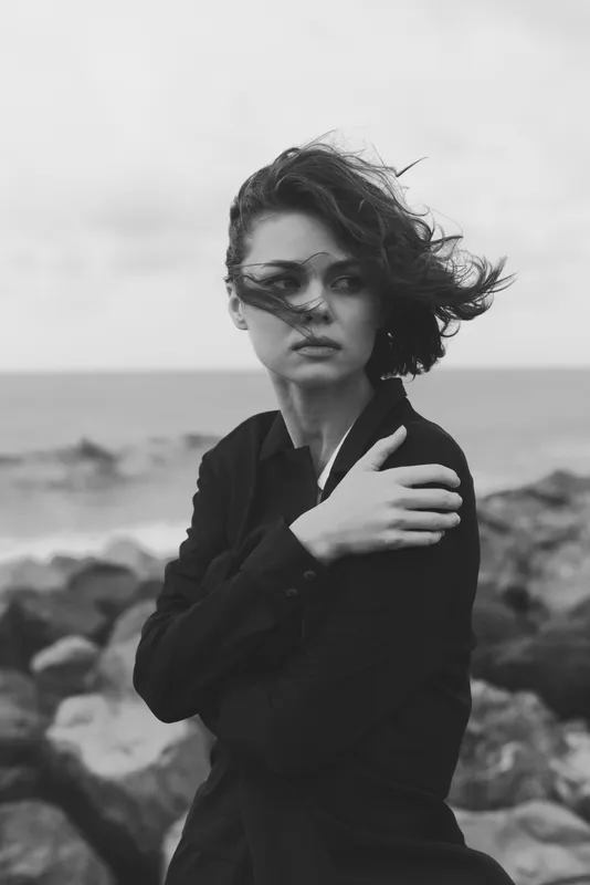 Woman with windswept hair by a rocky shoreline in monochrome, moody outdoor scene, ocean breeze, stylish coat, thoughtful pose, dramatic sky, wind and sea for fashion or editorial use