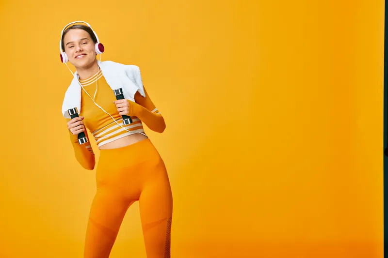 Female fitness athlete in bright orange activewear smiles in a studio, holding dumbbells with a white cropped jacket and headphones, dynamic workout vibes against a bold orange backdrop.