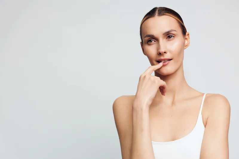 A thoughtful woman in a white tank top touches her lips with her finger while looking into the distance. The neutral background emphasizes her natural expression and skin tone.