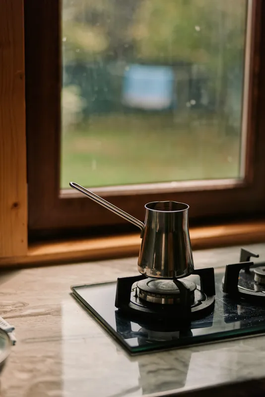 Kettle on a gas stove boils in a cozy kitchen by a wooden frame window, metal cookware and warm light creating a homely, calming cooking scene.