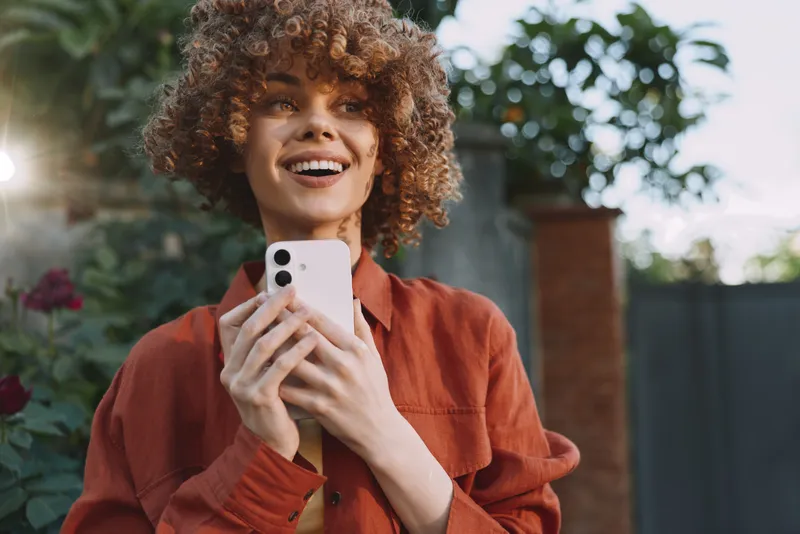 Happy woman lifestyle portrait outdoors, smiling while holding smartphone, casual red jacket, natural light, candid moment, modern urban vibe, carefree expression, authentic mood