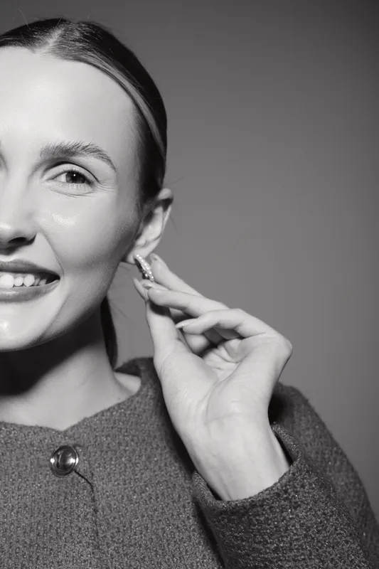 Smiling Caucasian woman with fair skin adjusts earring, wearing textured sweater against plain background. Concept of fashion and accessories.
