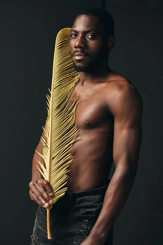 African man with dark skin holding large golden palm leaf in creative conceptual visual style. Portrait of shirtless male model with artistic expression on black background.