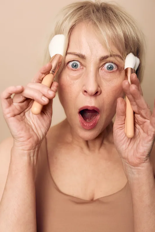 Surprised mature woman with wide blue eyes holding two makeup brushes near face. Concept of beauty routine, skincare, cosmetics application, and natural aging with expressive emotion.