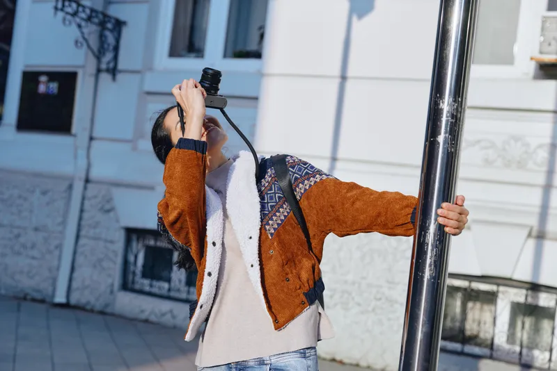 Outdoor fashion scene with an individual wearing a warm orange brown and cream jacket, posing with a selfie stick beside a light facade and sidewalk