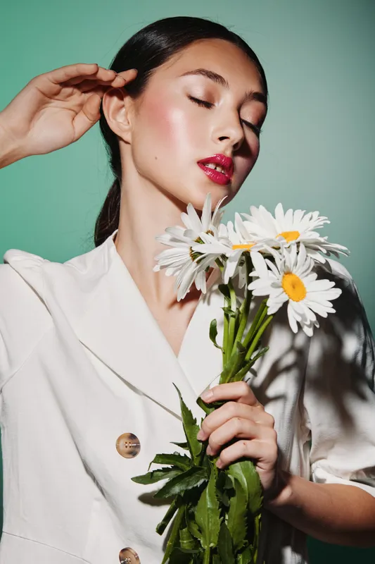 Elegant woman with flawless skin and red lips holding a bouquet of white daisies. She wears a beige jacket and poses gracefully against a green background. Beauty and fashion concept.