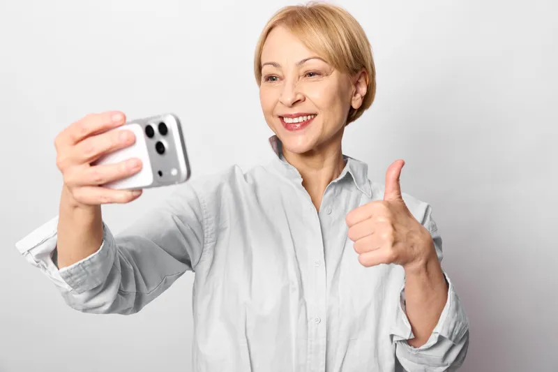 Woman selfie smartphone, senior smiling and giving thumbs up in a bright studio on white background, relaxed pose, casual shirt, soft light, for social posts