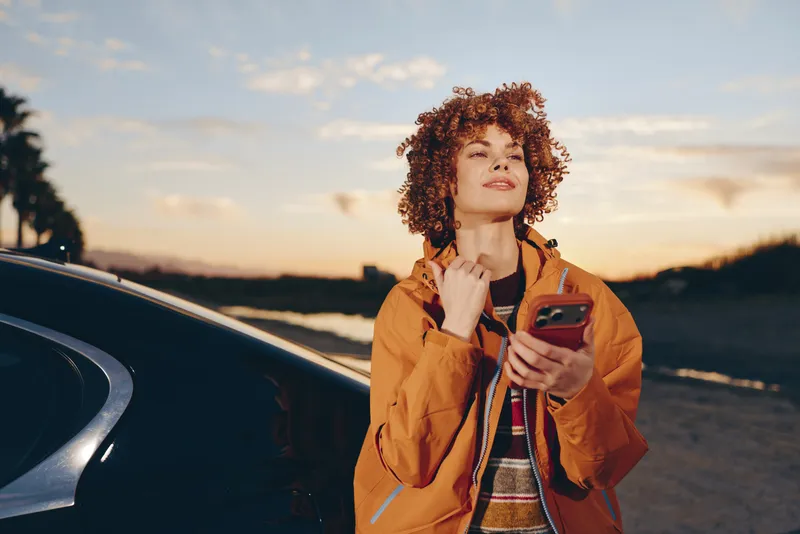 Woman lifestyle portrait outdoors near car at sunset, curly hair and warm smile, holding phone in hand, stylish casual jacket, soft light, open road background, relaxed travel mood