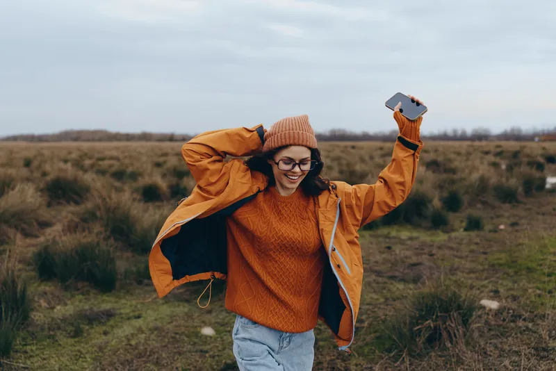 Woman outdoors in a field, lifestyle, orange jacket over knit sweater, autumn walking, smiling with arms up, windblown hair, cloudy sky, natural grass long horizon, soft breeze, relaxed pose