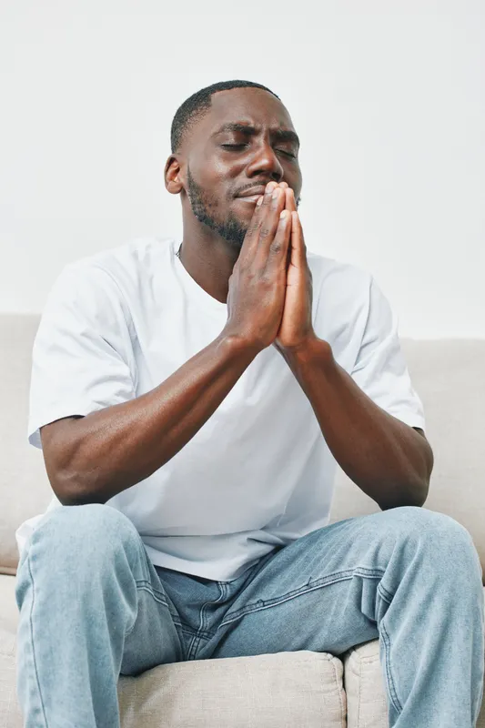 A contemplative African man in a white shirt sits on a couch with his hands clasped in prayer, conveying deep emotion and reflection in a minimalist setting