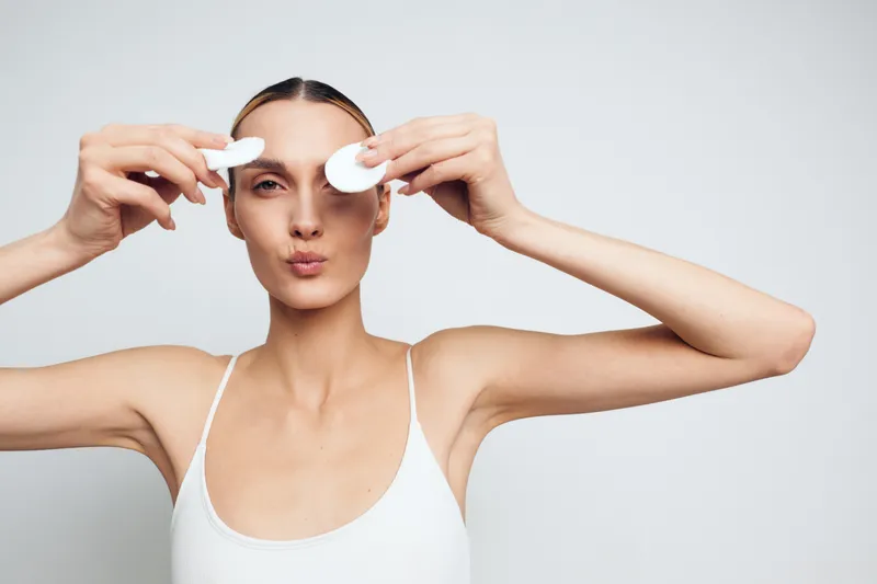Young woman with clear skin holding cotton pads near eyes, wearing white tank top on white background. Concept of skincare, beauty routine, and facial care with soft natural lighting.