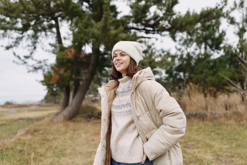 Woman in a warm beige coat and cream sweater stands in a windy park, wearing a knit beanie, casual winter fashion with trees and open field in the background.