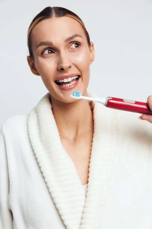 Young woman in white bathrobe brushes her teeth with electric toothbrush smiling and looking away in bright bathroom interior with white background and natural makeup.