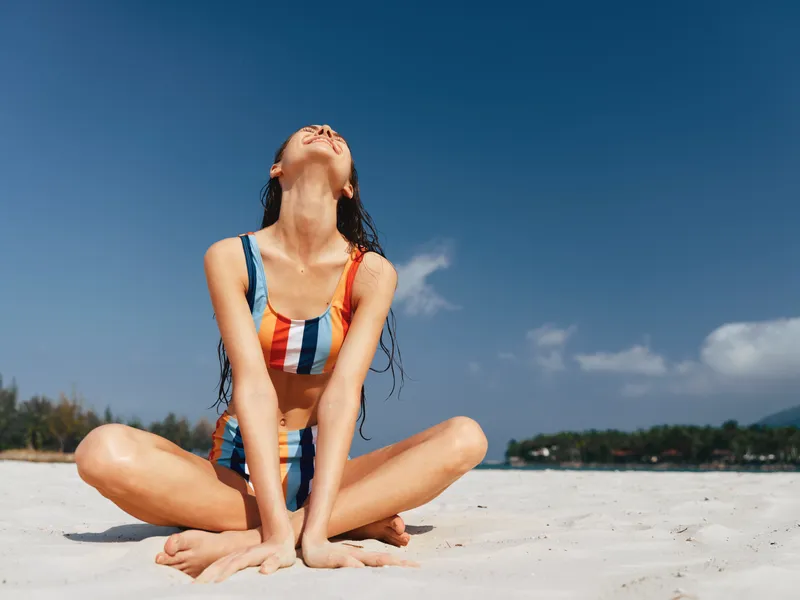 A young woman sits cross legged on white sand, wearing a striped bikini, back arched, head tilted toward the sun, capturing a moment of carefree beach relaxation.