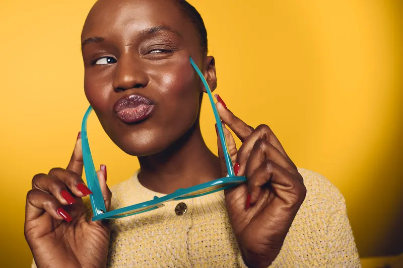 Playful dark skinned African woman holding blue sunglasses near face, winking and puckering lips. Yellow background, casual sweater. Concept for fun, summer, beauty, and fashion portrait.