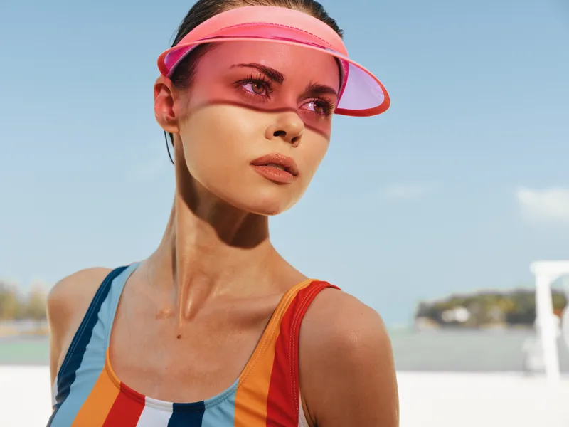 Portrait of a stylish young woman wearing a colorful striped swimsuit and pink visor under bright sun, her gaze toward the horizon conveying confidence and poise in the outdoors against a clear blue
