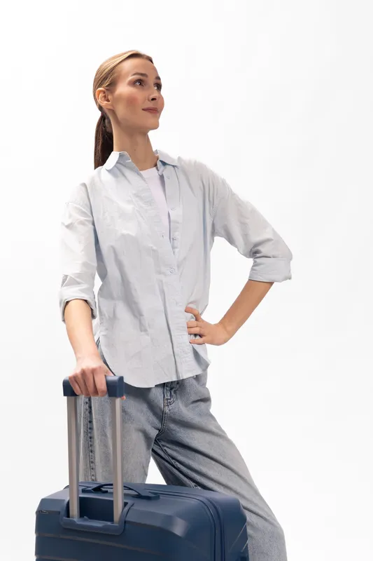 Confident young woman with long hair stands holding suitcase handle, looking away thoughtfully. Casual outfit and relaxed posture suggest travel, journey, or waiting concept with white background.