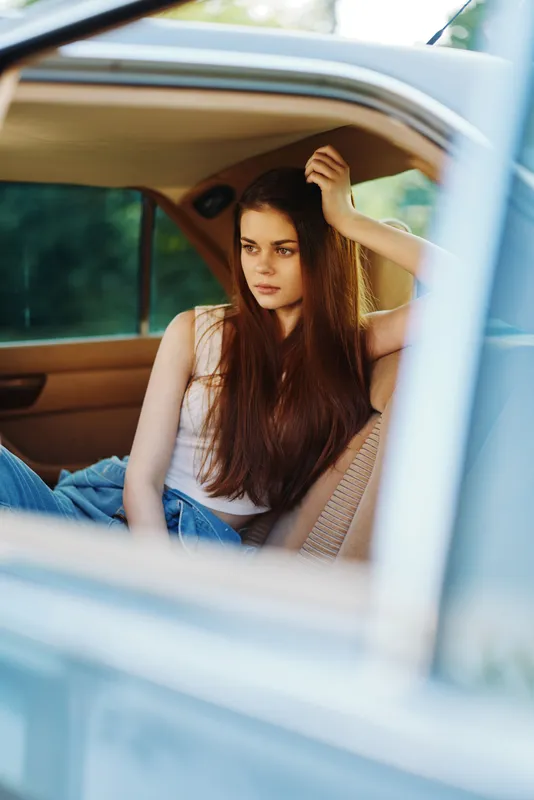 Young woman with long hair sitting in a vintage car, reflecting feelings of nostalgia and contemplation in a natural outdoor setting.
