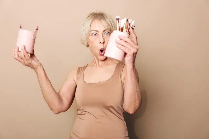 Surprised mature woman in beige tank top holding a pink container with makeup brushes against beige background. Concept of beauty tools, cosmetics, and unexpected discovery.