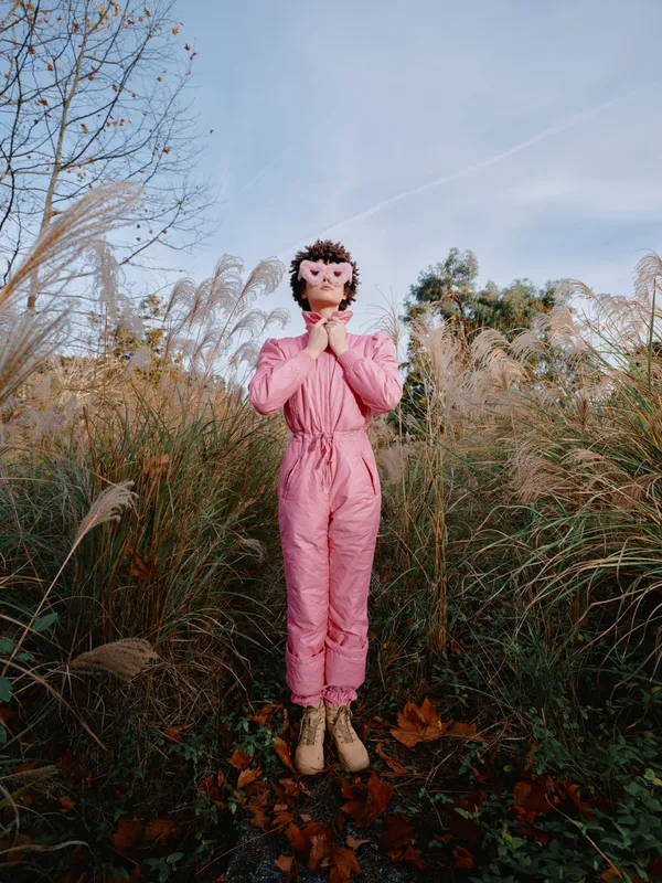 Outdoor scene with a person in a pink jumpsuit standing among tall grasses, hands in prayer, wearing a mask, autumnal setting, calm mysterious mood.