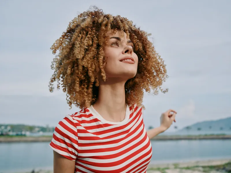 Portrait of a young woman with curly hair wearing a red and white striped shirt, standing outdoors beside a calm body of water under a clear blue sky.