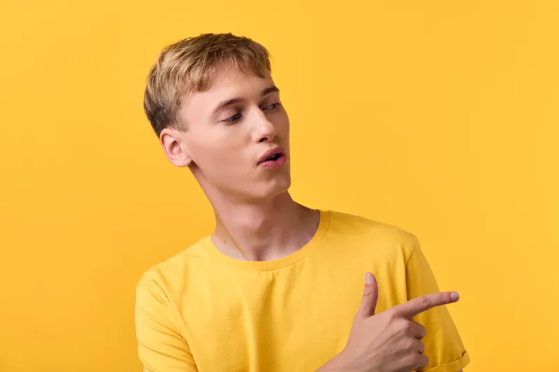 Young man in a yellow shirt points to the side against a bright yellow background, conveying direction, curiosity, and a casual mood for lifestyle and creative concept projects.