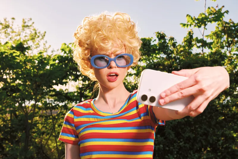 Woman with curly blond hair in a striped shirt and blue sunglasses takes a selfie outdoors, capturing a playful mood and retro color tones. Suitable for lifestyle and fashion imagery.
