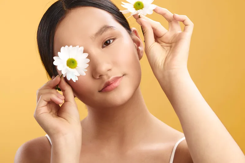 asian woman holding flowers near her face in a warm orange backdrop, soft portrait highlighting natural lighting, calm expression, and fresh beauty for a serene lifestyle moment.