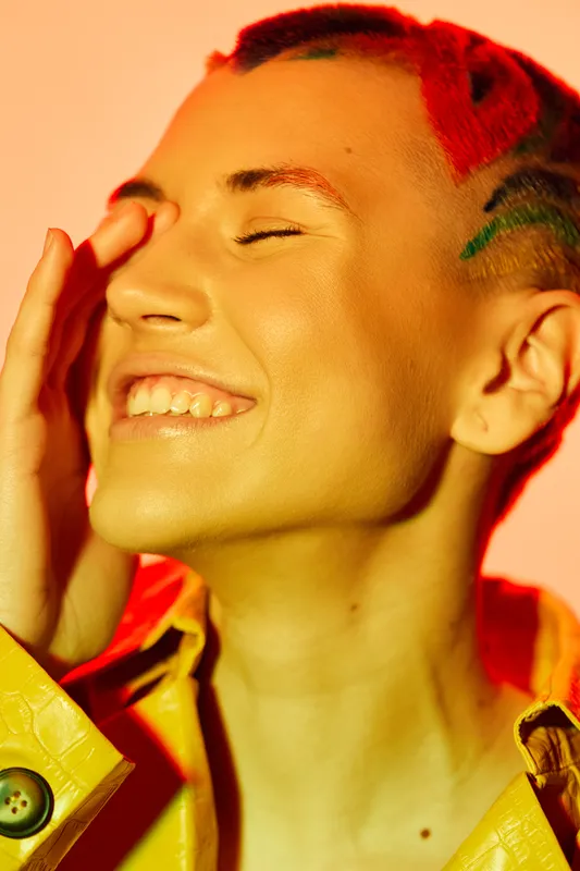 smiling woman with colorful shaved hair and yellow jacket enjoying vibrant warm light, expressive face, joyful emotion, creative style, close-up portrait