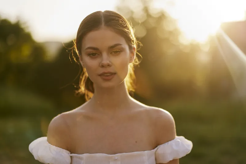 Young woman in a white dress with off shoulders, smiling softly in golden hour light, standing in a green garden Emphasizing beauty and tranquility in nature