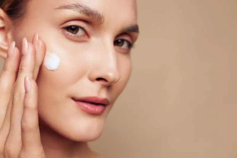 Woman skincare face cream beauty care routine moisturizer concept, close up of calm woman applying white cream under eye, smooth skin on warm beige background, copy space for ads.