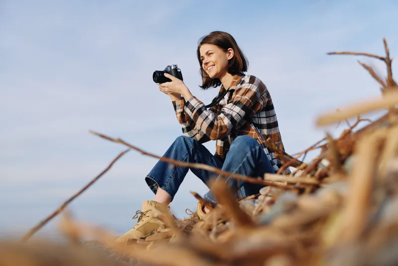 Photographer sits on a sandy dune, outdoors, with a camera ready, capturing nature and landscape moments as a woman in casual plaid shirt and jeans.