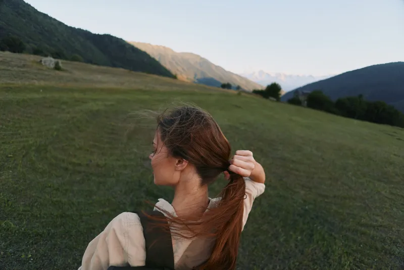 Young woman outdoors in an open field ties her hair back, enjoying a peaceful moment as a gentle breeze moves across the grass.