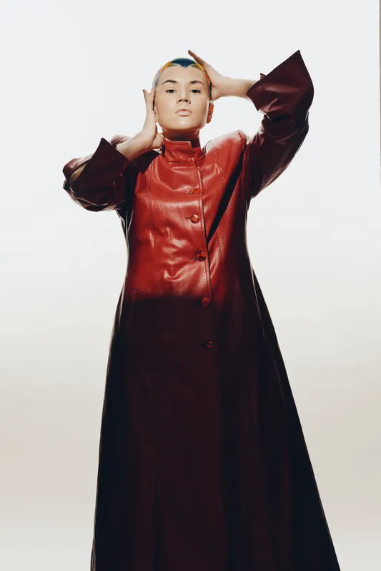 Young woman in long brown leather coat poses confidently against a minimal white background, displaying modern fashion and strong expression in studio lighting.